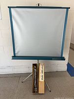 Front view of rolled-up projection screen fully extended with tripod legs; boxed tripod leaning against stand