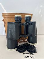 Front view of binoculars standing with lens caps and leather case behind