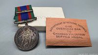 Medal with ribbon and bar beside original box and Defence slip
