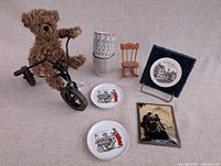 Group shot of all items: teddy on metal bicycle, two plates, two mugs, wooden chair, framed silhouette