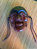 Full view of carved wooden mask showing color, carving detail, side cords, metal hanger