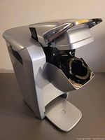 Front three-quarter view of silver Keurig brewer, lid open showing K-Cup chamber