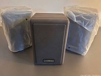 Front view of three Cambridge SoundWorks satellite speakers, two wrapped in plastic, showing size and grille design