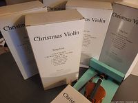Five sealed boxes labeled Christmas Violin with one open showing miniature violin
