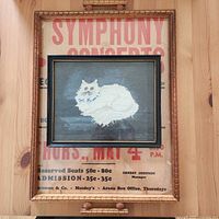 Needlework cat frame resting on wooden tray with concert poster