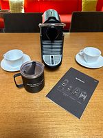 Front view of Breville espresso machine, two porcelain cup-and-saucer sets, black travel mug, and booklet on tabletop