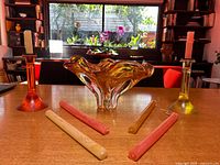 Front view of amber art glass bowl, two colored glass candlestick holders with candles, and loose tapers on table