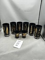 Eight black and gold Fred Press glasses arranged together, six tall highballs and two shorter lowballs