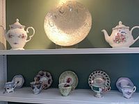 Shelf view of two floral teapots, pressed glass platter, five teacups and saucers