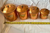 Four copper-tone canisters with lids lined up beside tape measure
