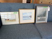 Three framed artworks on sofa: rural landscape, seascape, harbour scene
