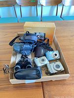 Box containing three Canon blue/silver digital cameras, three soft cases, Nikon binoculars, Canon retail box