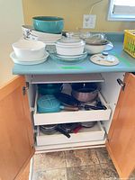 Countertop showing CorningWare casseroles, turquoise bowl, glass bowl, and cabinet with Danish pot, stainless cookware, fry pans