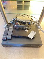 Stacked electronics showing Panasonic VCR, Samsung Blu-ray, remotes, headphones, power bar, cables