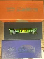 Three branded Pokémon TCG boxes: orange Sun & Moon, black Mega Evolution, purple Silver Tempest