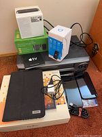 Stack of boxed electronics and loose devices showing Apple TV box, Doro phone box, blue-white device box, DVD player, tablet box, folio, smartphones and cables