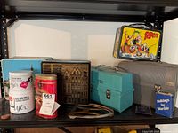 Shelf view showing Luxuria and Cadbury tins, Amsterdam tin, turquoise Thermos case, Popeye lunchbox, grey Thermos case, Wardair booklet, Souvenir Montreal trivet