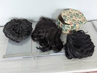 All three wigs and storage box on surface with rulers for scale