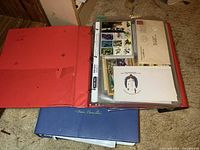 Red binder opened displaying stamp sheets, singles, first day covers in pocket pages, blue binder visible underneath