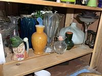Shelf showing multiple glass vases, satin glass bottle, purple compote, green bud vase, clear round vase, character mug
