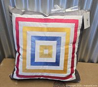 Front view of square pillow showing geometric concentric square design and attached tags