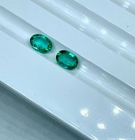 Two oval emeralds on white grooved tray, showing color and cut
