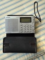 Front view of Grundig YB-300PE radio showing speaker, LCD, keypad and strap
