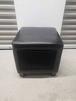 Front view of black faux leather ottoman showing one side pocket and wooden feet