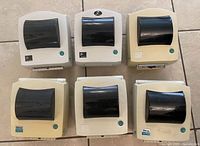 Six Zebra desktop label printers arranged in two rows, front view showing black lids and teal buttons