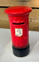Front view of red ceramic English mail box piggy bank showing coin slot and label