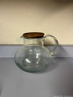 Front view of clear glass pitcher showcasing handle, bulbous base, wooden lid
