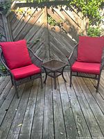 Full view of two patio chairs with red cushions and matching glass-topped side table on wooden deck