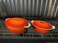 Two orange enameled cast iron Dutch ovens with lids on rack