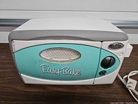 Front view of Easy-Bake teal aqua toy oven showing logo, vent window, timer knob, digital clock decal, attached power cord