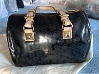 Front view of black patent Michael Kors satchel showing embossed MK pattern, beige handles, gold hardware