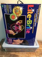 Retail box atop plastic bin, shows Lite-Brite branding