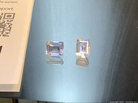 Two rectangular emerald-cut light rose quartz stones on reflective surface