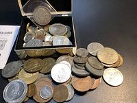 Open silver-tone box overflowing with assorted coins and tokens spilling onto table