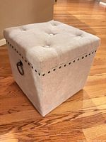 Closed ottoman showing light gray fabric, tufted lid, nailhead trim, ring handle