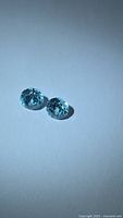 Two round brilliant cut blue-green zircons on white background