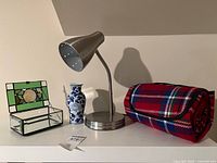 Group shot of stained glass box, porcelain vase, metal lamp and rolled plaid blanket on shelf