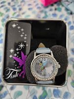 Watch in open tin showing dial, bezel, strap, and packaging