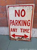 Full view of white metal NO PARKING ANY TIME sign with arrows, leaning against brick wall