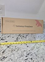 sealed box labeled Indoor/Outdoor Tabletop Fireplace with tape measure showing length