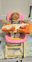 Doll seated in pink high chair with knit outfit pieces on tray