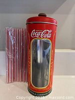 Tin Coca-Cola straw holder beside sealed pack of straws