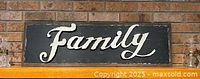 Full view of black wooden sign with raised metal word “Family” on brick backdrop