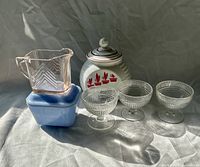 All six glass items grouped: grease jar, blue fridge container with lid, pink pitcher atop, three Hiawatha sherbets