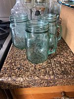 Three aqua-tinted mason jar drinking glasses with handles on countertop