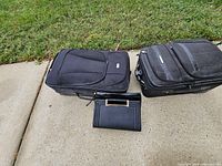 Two closed black rolling suitcases and black patent clutch on pavement